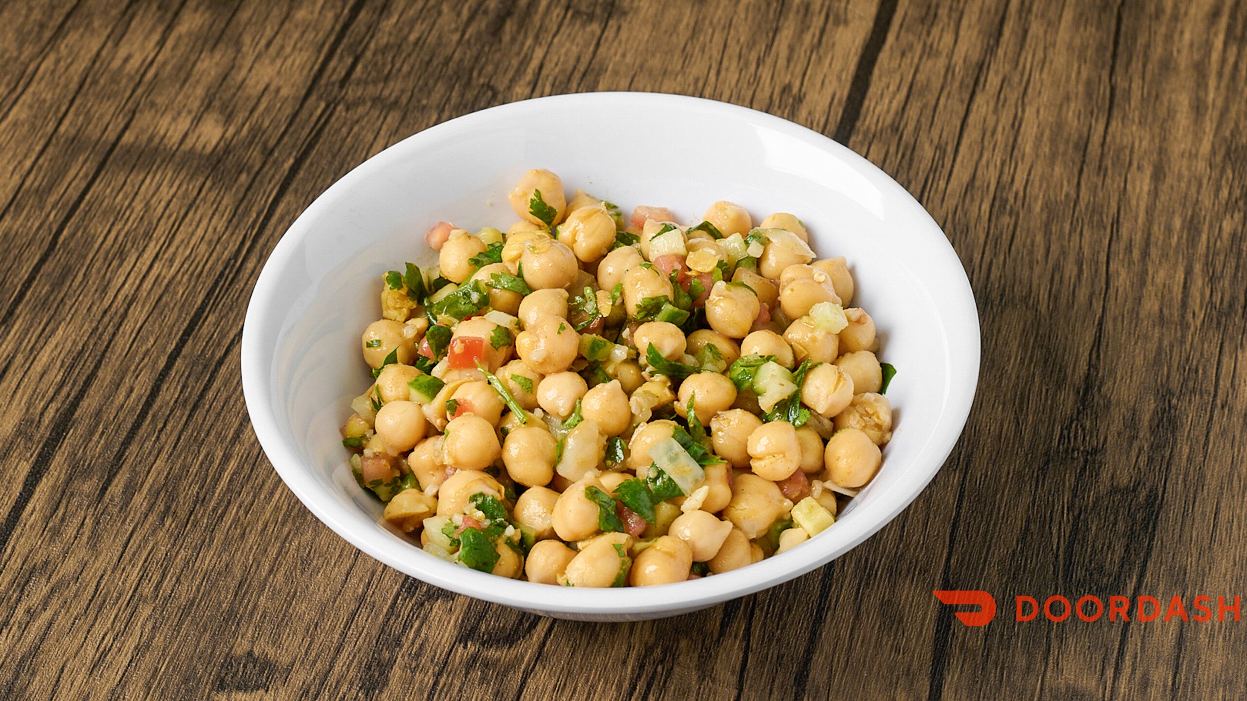 Chickpea salad in a white bowl on a wooden table with Doordash logo.