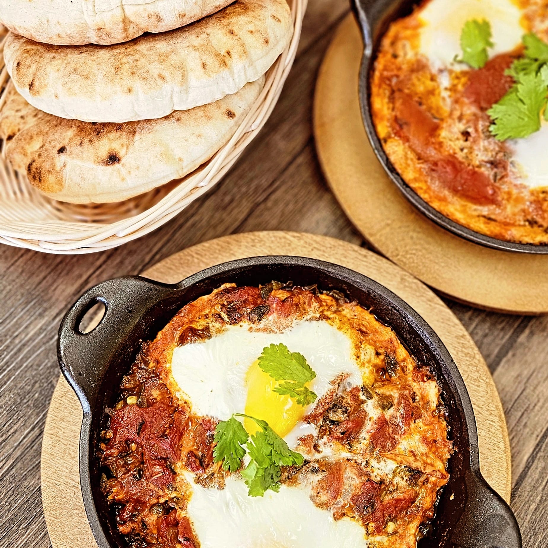 Egg dish with red sauce and herbs served in a cast iron skillet on a wooden table with flatbreads.