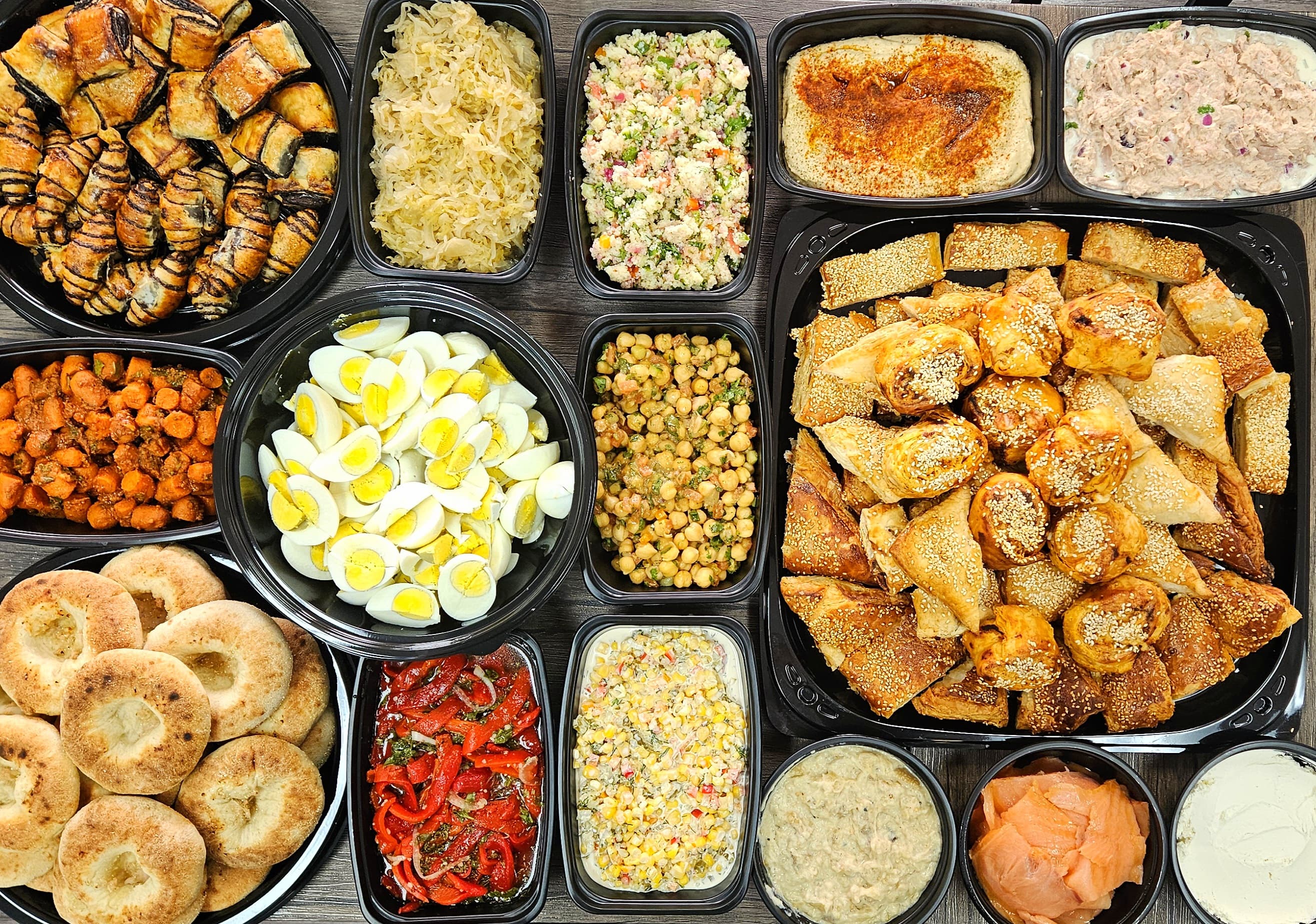 Assorted food items including burekas, salads, and dips in black catering containers.